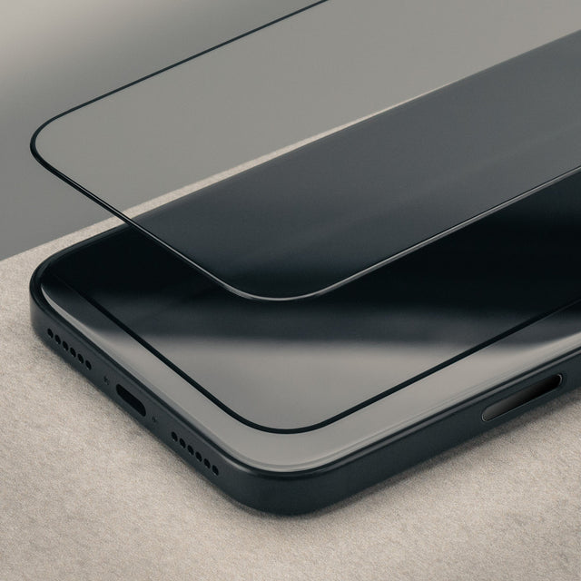 A close-up of a black smartphone with a transparent tempered glass screen protector partially lifted over the top surface, set on a light neutral textured surface.