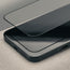 A close-up of a black smartphone with a transparent tempered glass screen protector partially lifted over the top surface, set on a light neutral textured surface.